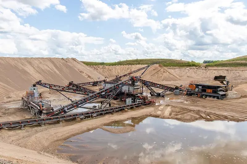 Large-scale sand and gravel quarry operation featuring multiple conveyor belt systems and processing equipment positioned among towering piles of extracted materials.