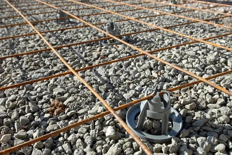Construction site showing steel rebar reinforcement grid laid out over a bed of gravel aggregate. Gray plastic spacers are positioned at grid intersections to maintain proper spacing and elevation of the rebar before concrete pouring.
