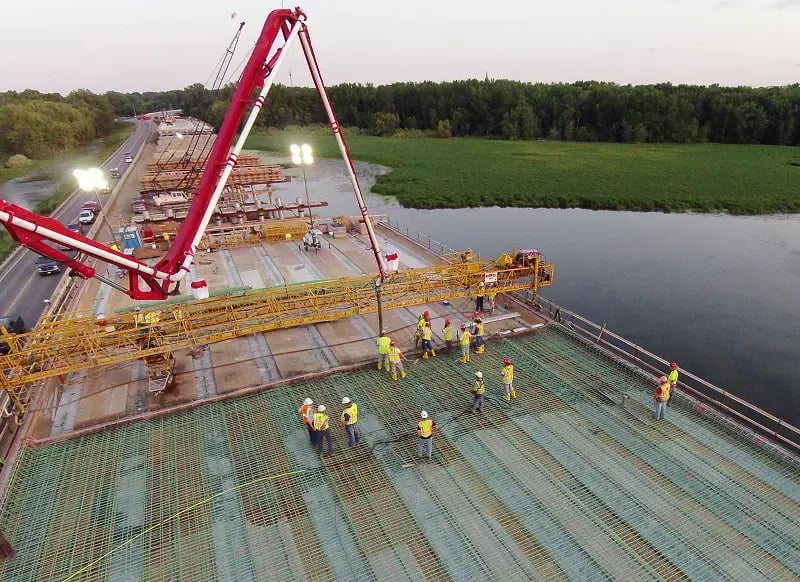 Aerial view of construction crew pouring concrete for bridge deck using red concrete pump truck with extended boom over water