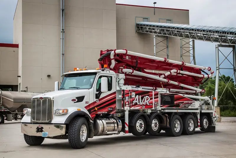 White concrete pump truck with red boom positioned at concrete plant facility with storage silos and conveyor system