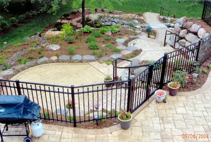 Residential backyard featuring curved decorative concrete pathways through landscaped rock garden with black metal fencing and plantings