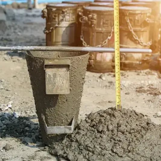 A concrete slump test setup on a construction site, showing a metal slump cone beside a freshly poured mound of concrete with a measuring tape to assess consistency.