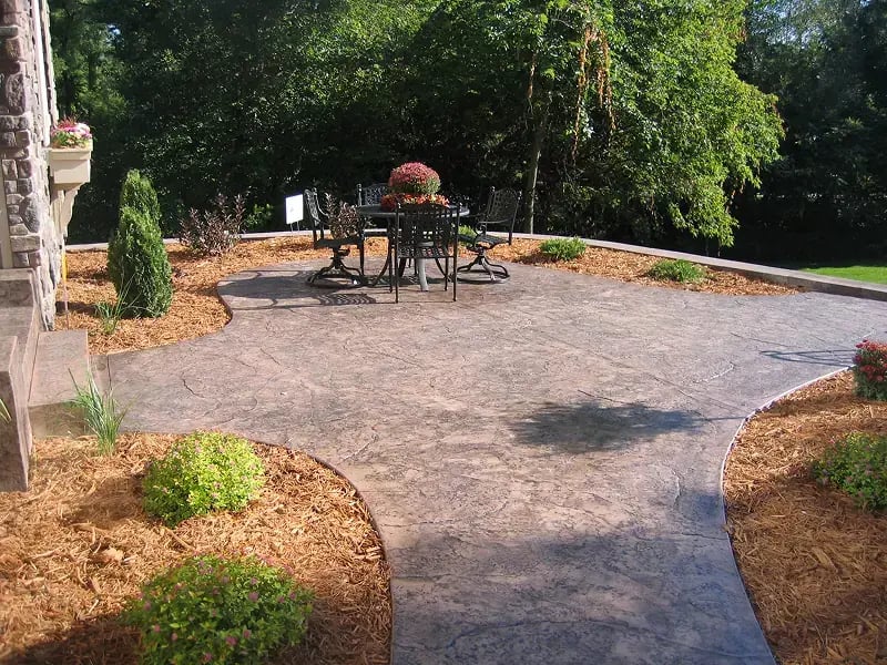 Curved stamped concrete patio with dark decorative pattern, furnished with black metal outdoor dining furniture. The patio is surrounded by landscaped beds with fresh mulch and plantings, adjacent to a brick home. Tree shadows create dappled light across the surface.