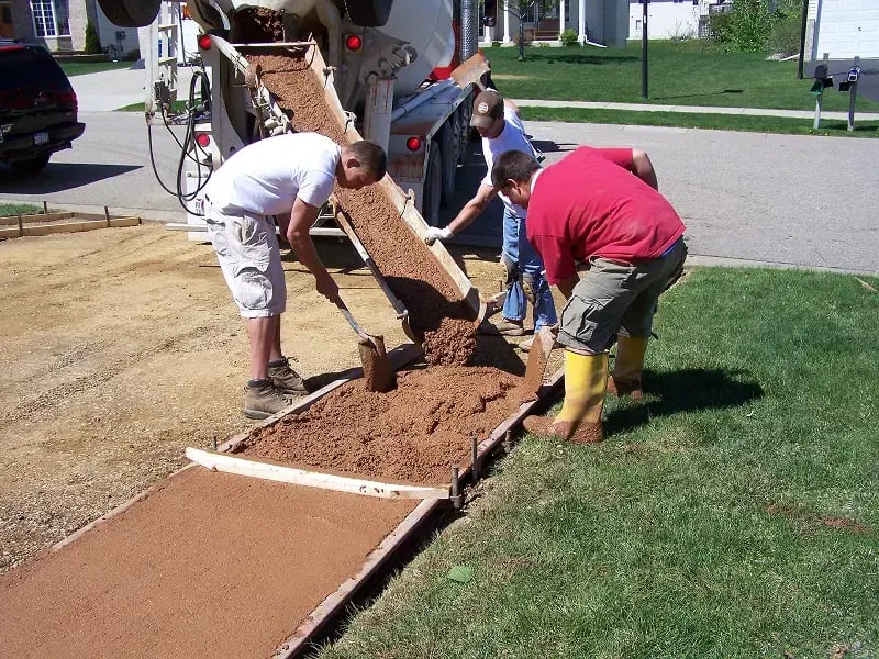 Three construction workers in a residential neighborhood pour and level concrete from a mixer truck into wooden forms for a driveway or walkway. The workers use tools to spread and smooth the wet concrete while the truck's chute directs the pour into the prepared forms.