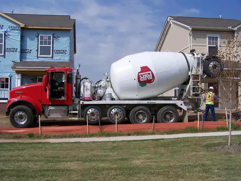 AVR Concrete mixer truck delivering concrete at a residential development site, with new homes under construction and a worker guiding the pour.