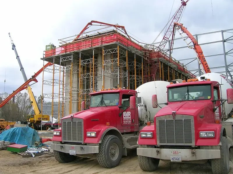 Two red AVR Concrete mixer trucks parked in front of a large commercial construction site with scaffolding, cranes, and concrete pumps in action.