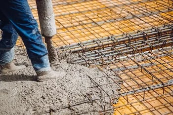 A construction worker pours wet concrete over a reinforced steel rebar grid, preparing the foundation of a building.