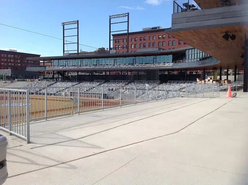 View of a modern baseball stadium under construction with installed seating, glass press boxes, and wide concrete walkways featuring accessibility ramps and metal railings.