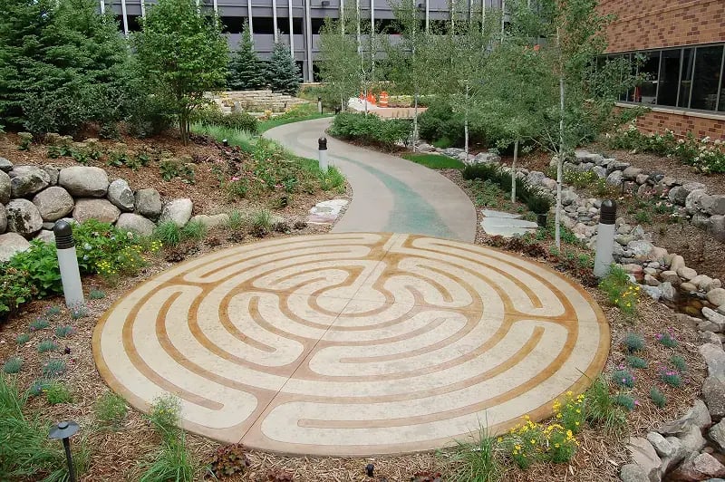 Circular concrete labyrinth design embedded in a landscaped garden path surrounded by flowers, rocks, and trees, creating a peaceful outdoor space.
