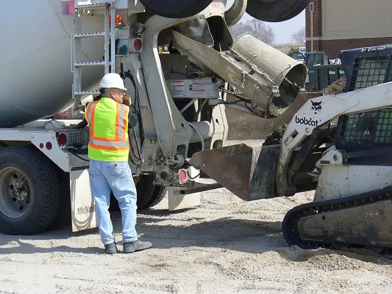A construction worker in a safety vest and helmet stands next to a cement truck and a Bobcat loader at a construction site.