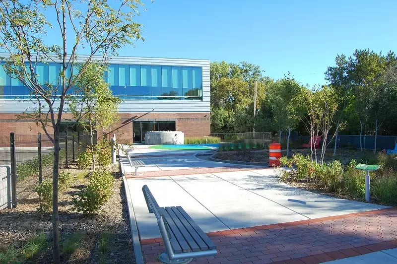 Landscaped courtyard outside a modern office building featuring young trees, benches, and clean concrete walkways bordered by brick pavers and greenery.