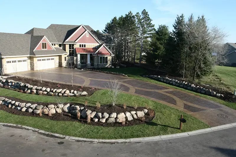 A large suburban house with multiple garages, a circular patterned concrete driveway, landscaped stone borders, and trimmed lawns surrounded by trees.
