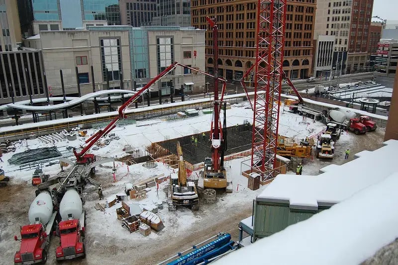 A construction site shows concrete being poured into a large foundation using multiple pump trucks and red booms. The active concrete pour and winter conditions highlight the challenges of cold-weather construction in the Twin Cities.