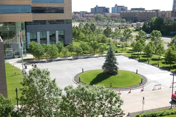 Aerial view of large concrete plaza with circular landscaped island featuring mature evergreen tree, surrounded by modern commercial buildings and urban streetscape