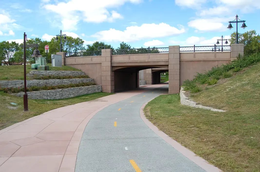 Concrete bridge underpass with decorative stone masonry retaining walls, colored concrete pathway, and ornamental lighting in park-like setting